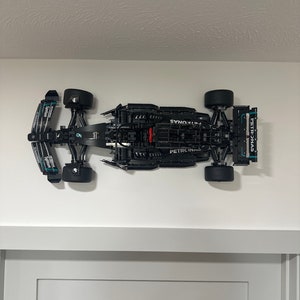 Puede incluir: Un modelo de coche de carreras de Fórmula 1 Lego negro con los logotipos de Petronas y Synops en el lateral. El coche está montado en una pared blanca.