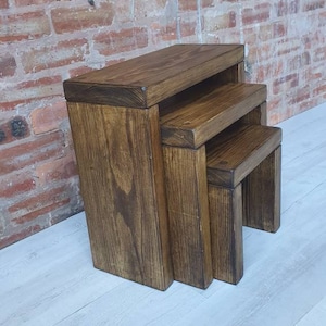 May include: A set of three wooden nesting tables in a dark brown finish. The tables are made of solid wood and have a simple, rustic design.