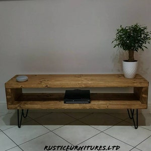 May include: A rustic wooden TV stand with a shelf and hairpin legs. The stand is made of reclaimed wood and has a natural finish. The stand is 47 inches wide, 16 inches deep, and 18 inches tall.