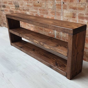 May include: A dark stained wooden shelf unit with two lower shelves. The rectangular unit has a modern design. The wood grain is visible. The unit is against a brick wall.
