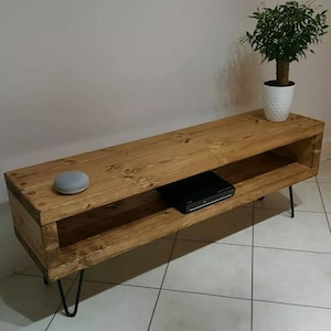 May include: A rustic wooden TV stand with a shelf and hairpin legs. The stand is stained a dark brown color and has a natural wood grain. It is sitting on a white tiled floor.