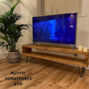 May include: A rustic wooden TV stand with two shelves and hairpin legs. The stand is made of reclaimed wood and has a natural finish. A flat screen TV is sitting on top of the stand.