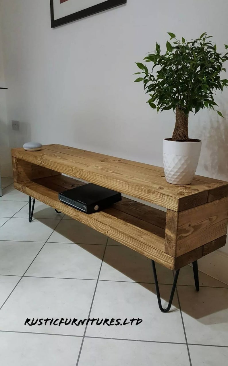 May include: A rustic wooden TV stand with a shelf, featuring a natural wood finish and black hairpin legs. The stand is displayed on a tiled floor with a potted plant and a small speaker on top.
