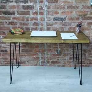 May include: A wooden desk with a light brown finish and black hairpin legs. A laptop computer, a stack of books, and a piece of paper are on the desk.