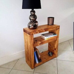 May include: A rustic wooden side table with two shelves. The table is stained a dark brown colour and has a distressed finish. A black lamp with a round, silver base sits on the top shelf. There are books and a small, round object on the lower shelf. The text "RUSTICFURNITURES.LTD" is visible at the bottom of the image.
