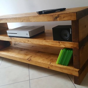 May include: A rustic wooden entertainment unit with two shelves. The top shelf features a games console and a remote control. The bottom shelf has a stack of books and a stack of video games. The unit is made from natural wood with a dark brown stain.