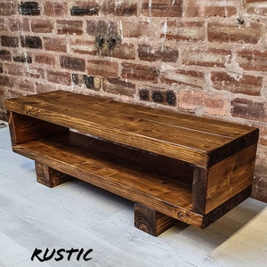May include: A rustic wooden TV stand with a shelf, stained in a dark brown color. The stand has a rectangular shape with four legs and a cutout on the back. The stand is sitting on a light wood floor.