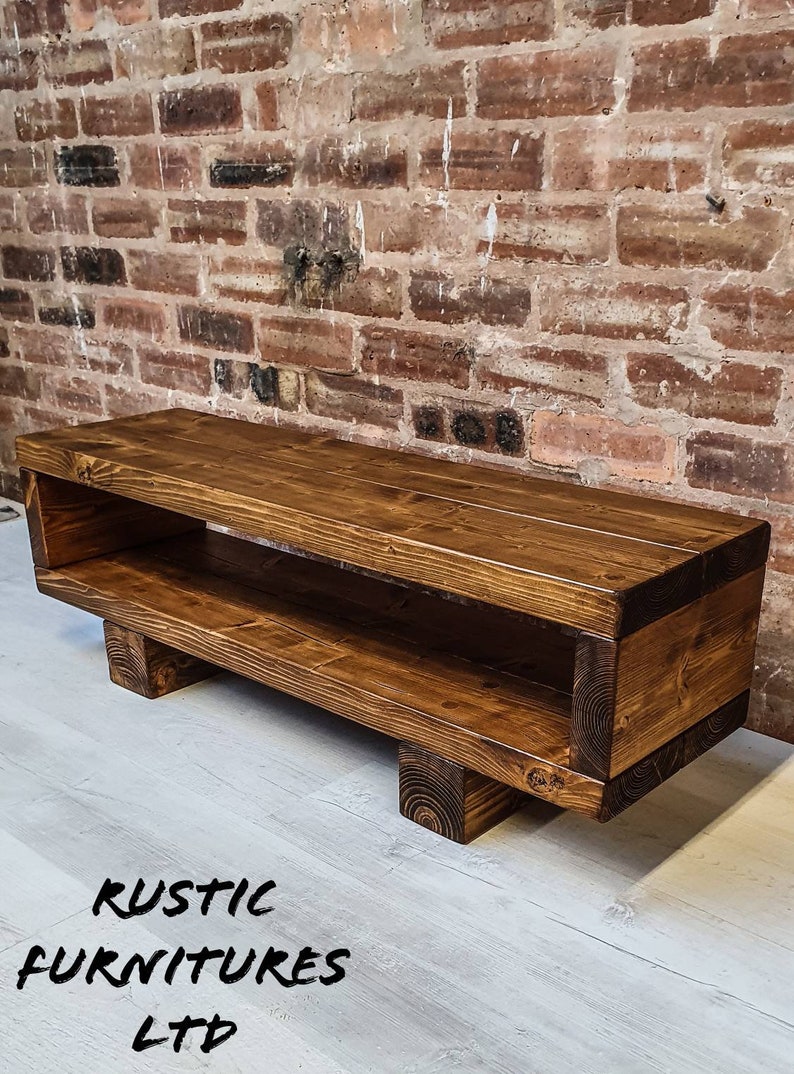 May include: A rustic wooden TV stand with a shelf, stained in a dark brown color. The stand has a rectangular shape with four legs and a cutout on the back. The stand is sitting on a light wood floor.