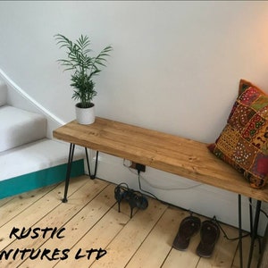 May include: A rustic wooden bench with black hairpin legs. The bench is decorated with a colourful patterned cushion and is placed in a hallway with a staircase in the background. The text "RUSTIC FURNITURES LTD" is visible in the bottom left corner of the image.
