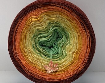 May include: A skein of yarn in a gradient of orange, yellow, green, and brown colours. The yarn is wound into a ball shape and has a small leaf charm attached.