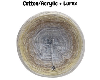 May include: A skein of yarn in a gradient of beige, light brown, and silver with a silver lurex thread. The yarn is wound into a ball shape and the label reads "Cotton/Acrylic + Lurex".