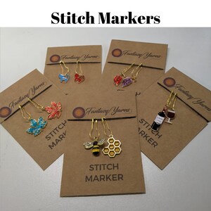 May include: Five stitch markers with different charms on brown cards. The charms include a bee and honeycomb, a wine bottle and glass, a maple leaf, a red leaf, and a blue bird. The cards have the text "Fantasy Yarns" and "Stitch Marker" on them.