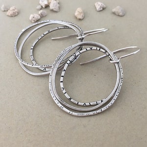 May include: Silver hoop earrings. Each earring has three textured, interconnected rings with etched patterns. The earrings hang from simple ear wires. The image shows the earrings on a neutral surface, with small stones scattered around.