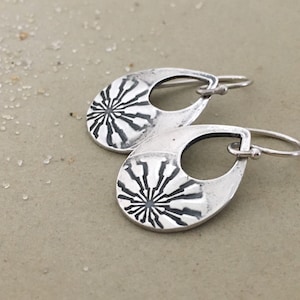 May include: A pair of silver dangle earrings with a geometric design. The earrings are shaped like teardrops with a circular cutout in the center. The design features a radiating pattern of lines.