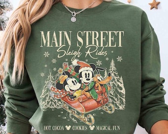 May include: Green sweatshirt with a vintage-style graphic of Mickey Mouse, Minnie Mouse, and Goofy riding in a sleigh. The text "Main Street Sleigh Rides" is above the graphic, and "Hot Cocoa Cookies Magical Fun" is below.