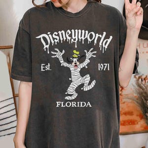 May include: Dark grey t-shirt with a Disney World design. The text reads "Disneyworld", "Est. 1971", and "Florida". The graphic depicts Goofy as a mummy. The shirt is being worn by a person.
