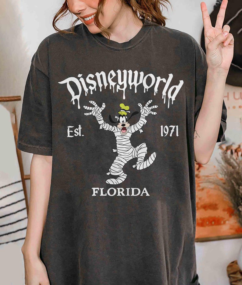 May include: Dark grey t-shirt with a Disney World design. The text reads "Disneyworld", "Est. 1971", and "Florida". The graphic depicts Goofy as a mummy. The shirt is being worn by a person.