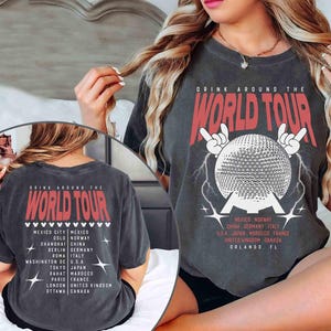 May include: Dark gray t-shirt with the text "DRINK AROUND THE WORLD TOUR" in red. The front features a globe design with lightning bolts and the text "MEXICO NORWAY CHINA GERMANY ITALY USA JAPAN MOROCCO FRANCE UNITED KINGDOM CANADA ORLANDO, FL".