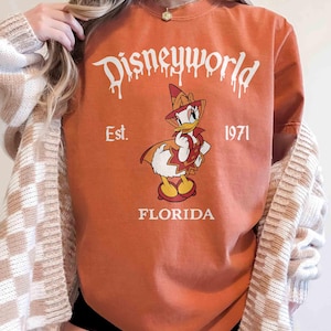 May include: Orange t-shirt with a Disneyworld graphic featuring Donald Duck in a pilgrim outfit. The text "Disneyworld" is above the duck, with "Est. 1971" and "Florida" below. The shirt is a crew neck.