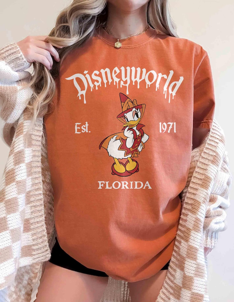May include: Orange t-shirt with a Disneyworld graphic featuring Donald Duck in a pilgrim outfit. The text "Disneyworld" is above the duck, with "Est. 1971" and "Florida" below. The shirt is a crew neck.