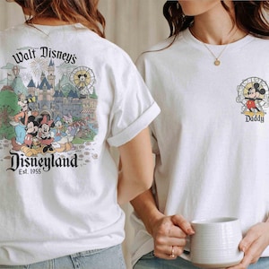 May include: White t-shirts featuring Disney characters. One shirt has a Disneyland graphic on the back, with Mickey, Minnie, Donald, and Goofy. The other shirt has a small Mickey Mouse graphic with the word "Daddy".