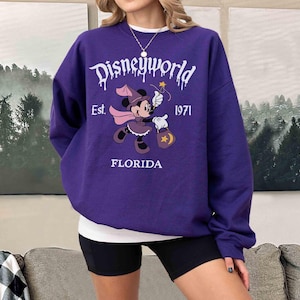 May include: A purple sweatshirt with the text "Disneyworld" in white dripping font. The sweatshirt features a graphic of Minnie Mouse in a witch costume, holding a wand and a bag. The text "Est. 1971 FLORIDA" is also printed on the sweatshirt.