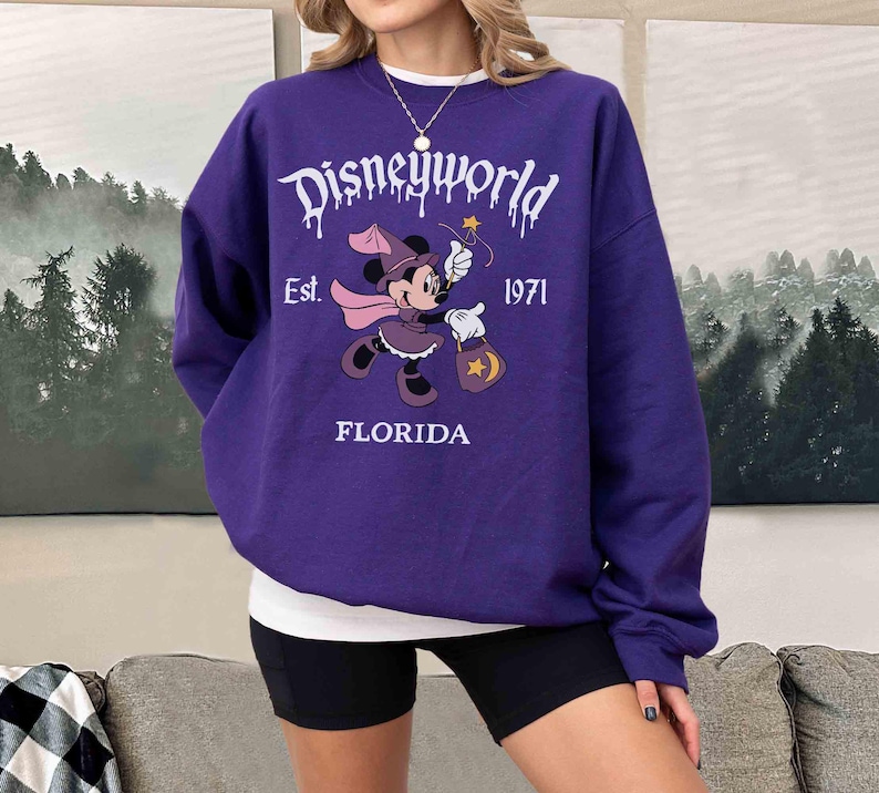 May include: A purple sweatshirt with the text "Disneyworld" in white dripping font. The sweatshirt features a graphic of Minnie Mouse in a witch costume, holding a wand and a bag. The text "Est. 1971 FLORIDA" is also printed on the sweatshirt.