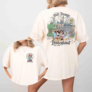 May include: Cream-colored t-shirt featuring a Disneyland graphic on the back with Mickey Mouse, Minnie Mouse, and other Disney characters. The front of the shirt has a small graphic of Minnie Mouse with the word "Mommy".