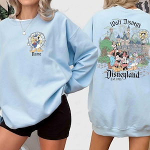 May include: Light blue sweatshirt featuring Donald Duck in front and a Disneyland graphic on the back. The front design includes Donald Duck in a Ferris wheel with the word "Name" below. The back design showcases Disney characters and the Disneyland castle.