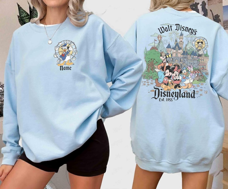May include: Light blue sweatshirt featuring Donald Duck in front and a Disneyland graphic on the back. The front design includes Donald Duck in a Ferris wheel with the word "Name" below. The back design showcases Disney characters and the Disneyland castle.