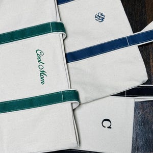 May include: Three canvas tote bags with different colored stripes and embroidered initials. The first bag has green stripes and says "Cool Mom". The second bag has blue stripes and the initials "SRM". The third bag has black stripes and the initial "C".