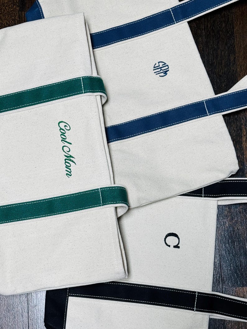 May include: Three canvas tote bags with different colored stripes and embroidered initials. The first bag has green stripes and says "Cool Mom". The second bag has blue stripes and the initials "SRM". The third bag has black stripes and the initial "C".