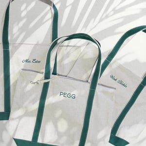 May include: Three white canvas tote bags with green straps. The bags are personalized with the names "Mrs. Estes", "Work. Bikini", and "PEGG".