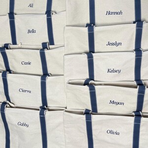 May include: Eight white canvas tote bags with navy blue handles and trim. Each bag has a name embroidered on the front in white thread: Ali, Bella, Cassie, Ciarra, Gabby, Hannah, Jesslyn, Kelsey, Megan, and Olivia.