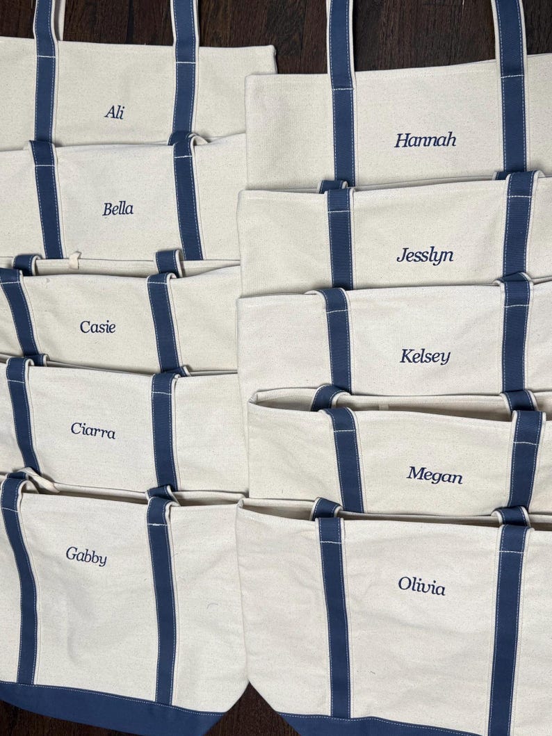May include: Eight white canvas tote bags with navy blue handles and trim. Each bag has a name embroidered on the front in white thread: Ali, Bella, Cassie, Ciarra, Gabby, Hannah, Jesslyn, Kelsey, Megan, and Olivia.