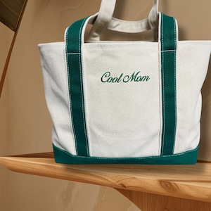May include: A white canvas tote bag with green trim and the words "Cool Mom" embroidered in green.