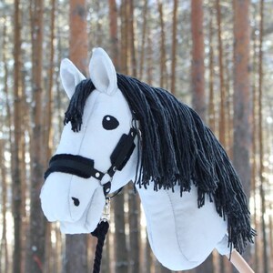 May include: A white hobby horse with a black mane and bridle. The toy horse has a black rope and is set against a blurred background of trees. The horse's head is detailed with black eyes and a black nose.