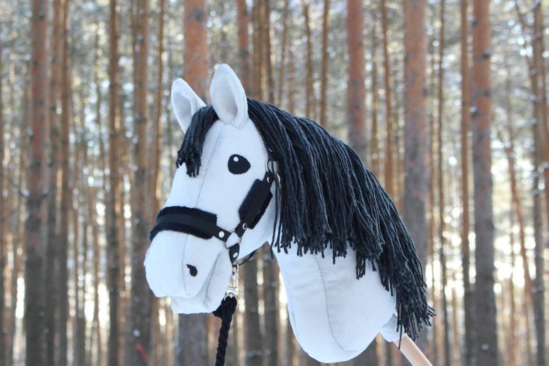 May include: A white hobby horse with a black mane and bridle. The toy horse has a black rope and is set against a blurred background of trees. The horse's head is detailed with black eyes and a black nose.