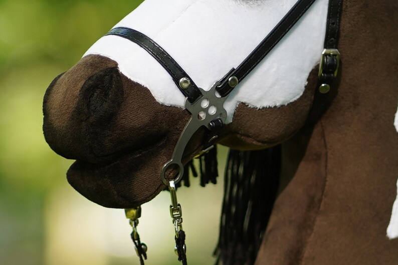 Puede incluir: Primer plano de una cabeza de caballo de peluche marr&oacute;n y blanco con una brida de cuero negro y una boquilla de plata. El caballo tiene una crin y una cola negras.
