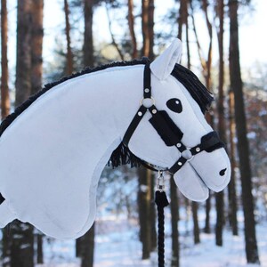 May include: A light blue hobby horse with a black mane and bridle. The toy horse has a wooden stick for a handle and is set against a snowy forest backdrop. The horse's bridle is black with gold-colored accents.