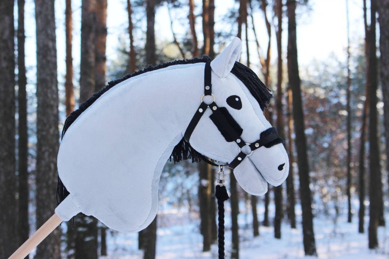 May include: A light blue hobby horse with a black mane and bridle. The toy horse has a wooden stick for a handle and is set against a snowy forest backdrop. The horse's bridle is black with gold-colored accents.