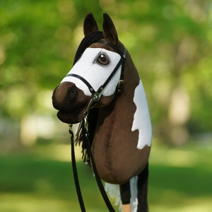 Puede incluir: Una cabeza de caballo pinto marr&oacute;n y blanco con una brida y riendas negras. El caballo es un juguete de peluche y est&aacute; de pie sobre un fondo verde de hierba.