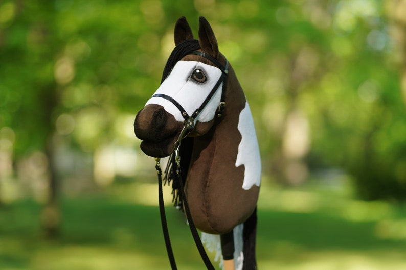 Puede incluir: Una cabeza de caballo pinto marr&oacute;n y blanco con una brida y riendas negras. El caballo es un juguete de peluche y est&aacute; de pie sobre un fondo verde de hierba.