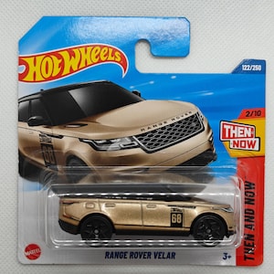 May include: A gold Hot Wheels toy car of a Range Rover Velar with black accents and the number 68 on the side. The car is on a blue and white card with the words "Then and Now" and "Range Rover Velar".