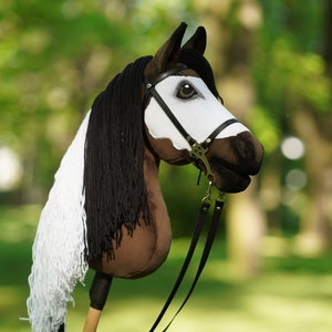 Puede incluir: Una cabeza de caballo marr&oacute;n y blanca con crin y cola negras, llevando una brida de cuero negro con bocado de plata. La cabeza del caballo est&aacute; unida a un palo de madera, y el fondo es verde borroso.