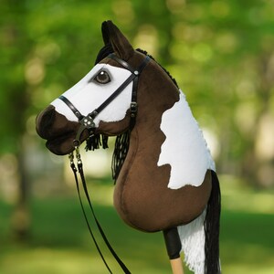 Puede incluir: Un caballo de palo pintado de marr&oacute;n y blanco con una brida y riendas negras. El caballo tiene una crin y una cola blancas.