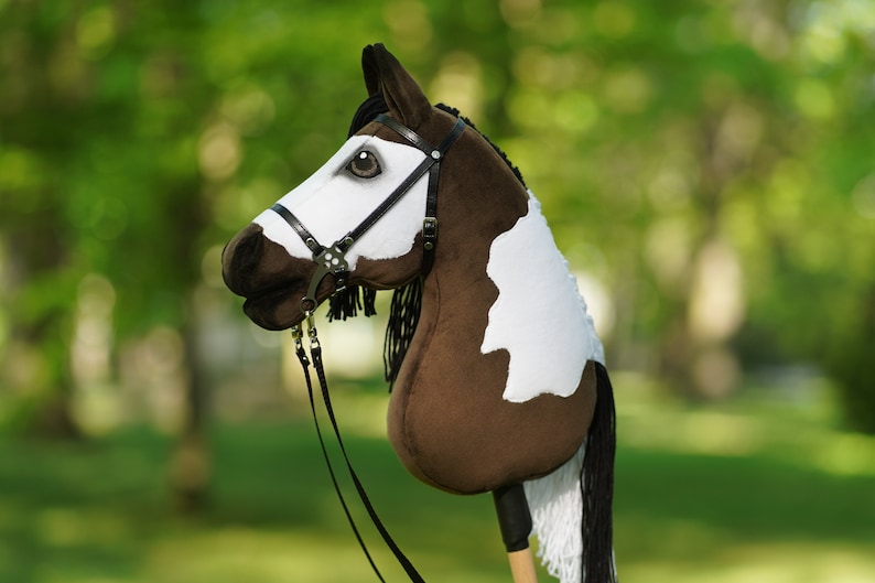 Puede incluir: Un caballo de palo pintado de marr&oacute;n y blanco con una brida y riendas negras. El caballo tiene una crin y una cola blancas.
