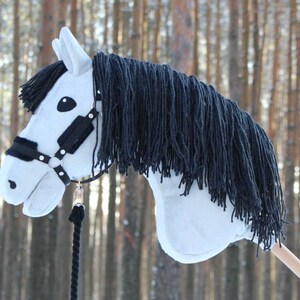 May include: A white hobby horse with a black mane and bridle. The toy horse has a light-colored wooden stick for a handle and a black rope. The horse's head is detailed with black eyes and a black bridle.