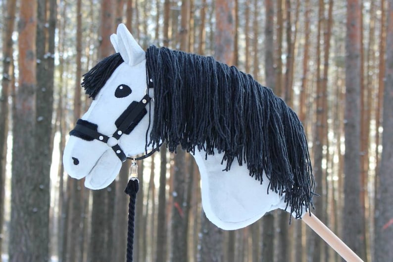 May include: A white hobby horse with a black mane and bridle. The toy horse has a light-colored wooden stick for a handle and a black rope. The horse's head is detailed with black eyes and a black bridle.