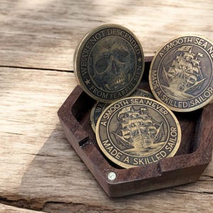 May include: Three antique brass coins with engraved designs and text. One coin features a skull, while the others depict a sailing ship. The coins are displayed in a small, dark wooden tray. The text on the coins reads "WE ARE NOT DESCENDED FROM FEARFUL MEN" and "A SMOOTH SEA NEVER MADE A SKILLED SAILOR."